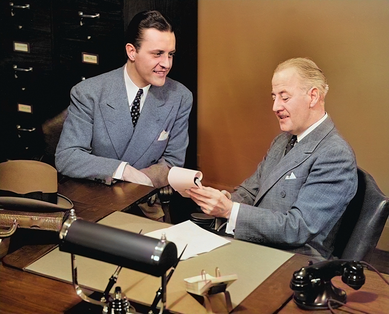 Colorized photograph of two men wearing suits seated at a desk