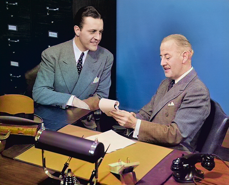 Colorized photograph of two men wearing suits seated at a desk next to blue wall