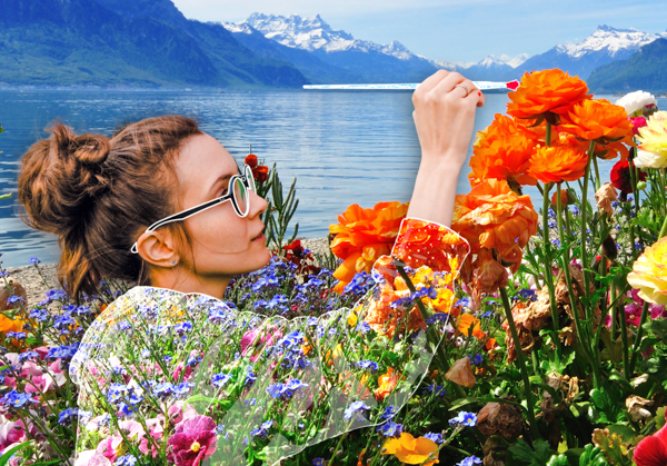 Altered photo of young woman with long brown hair in bun painting a photo of a flower with mountain and water in background. Pattern on her clothing merges with flowers in foreground.