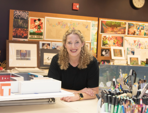 Woman with blonde curly hair sits at desk and looks directly at reader