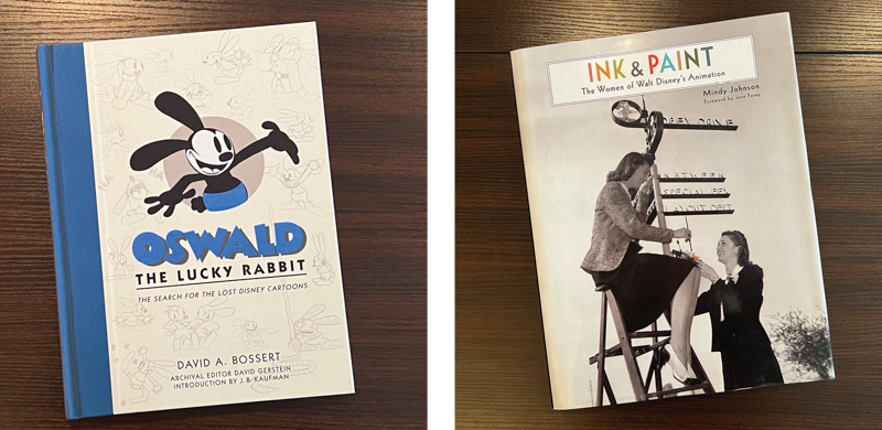 Two photos, each with a book tilted at an angle on a tabletop: Oswald the Lucky Rabbit, with black cartoon rabbit poking through a porthole against a background of pencil sketches, and Ink & Paint, a cover featuring two women, one sitting on the top of a ladder with a brush and another holding a palette for her, next to sign "Disney Drive".