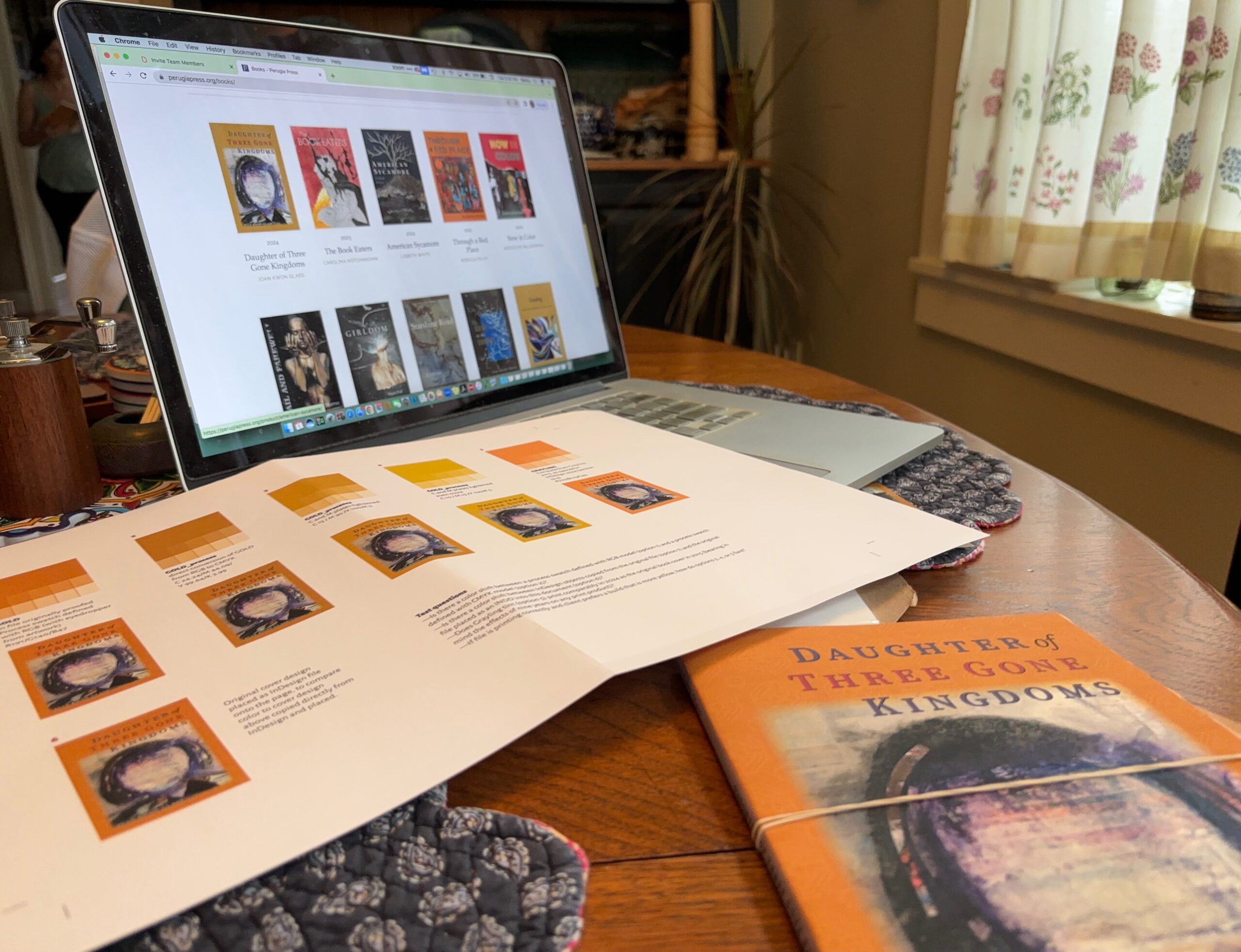 A laptop computer shows a website with book covers; atop the keyboard is the proof sheet that is the subject of this article and an unbound proof of the book whose cover is orange and not gold.