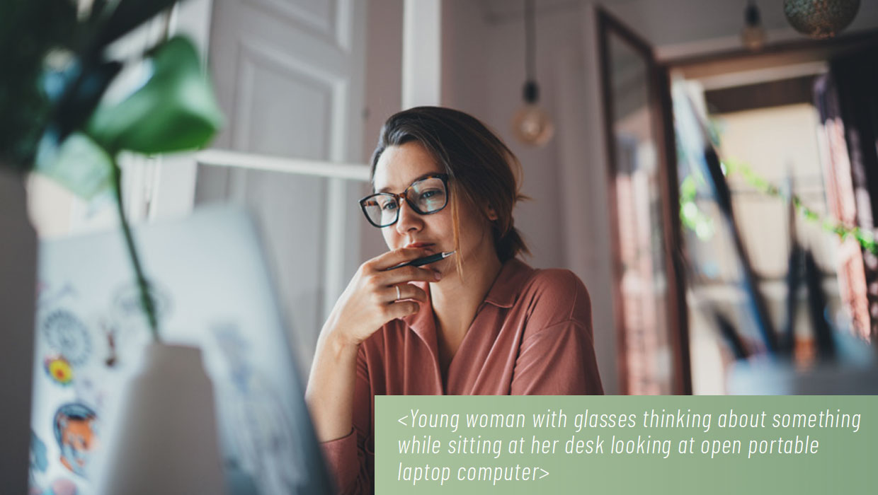 Young woman with glasses thinking about something while sitting at her desk looking at open portable laptop computer