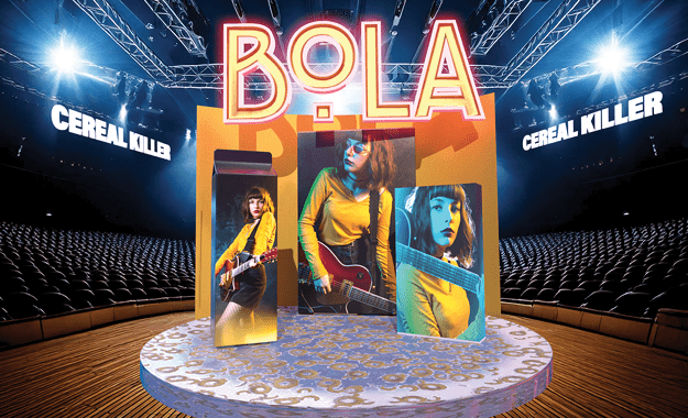 A simulated empty auditorium with extruded logo "BOLA" in center. "Cereal Killer" in extrabold sans serif caps on either side, at 1-point perspective. Three still photos of woman with guitar are centered on a platform with the cereal design, against a plain orange backdrop. All casts a dark shadow on the platform. The platform is on a brown wood stage. Two spotlights illuminate from behind from either side of the setup.